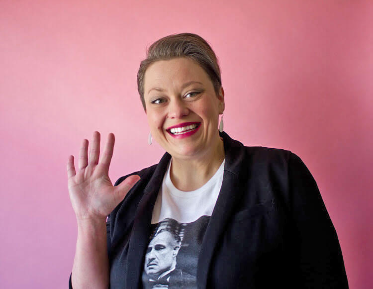 Melissa-Ann Dailey smiling and waving while standing in front of a pink background